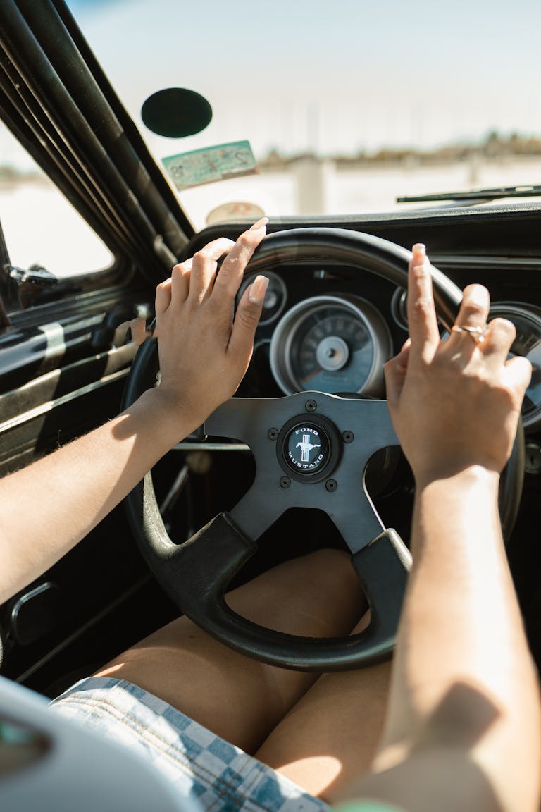 Hands on the steering wheel of a vintage Ford Mustang driving through Los Angeles on a sunny day.