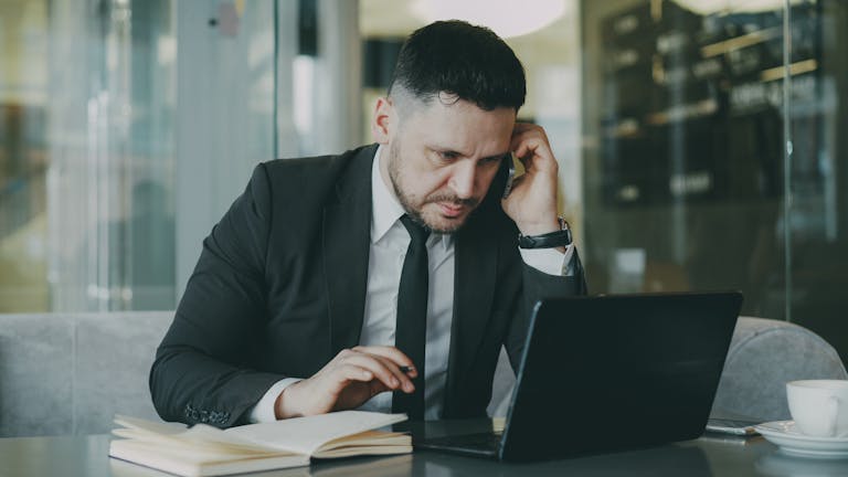 Focused businessman in suit multitasking on phone and laptop in modern office.