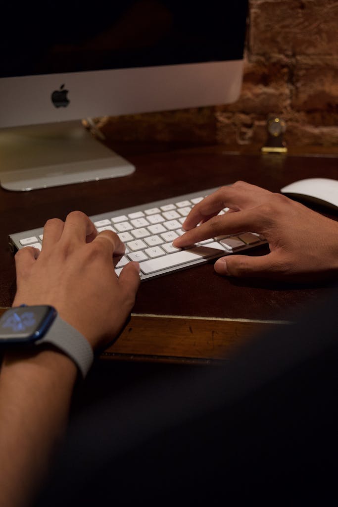 Hands typing on a desktop keyboard with a smartwatch visible, indoors.