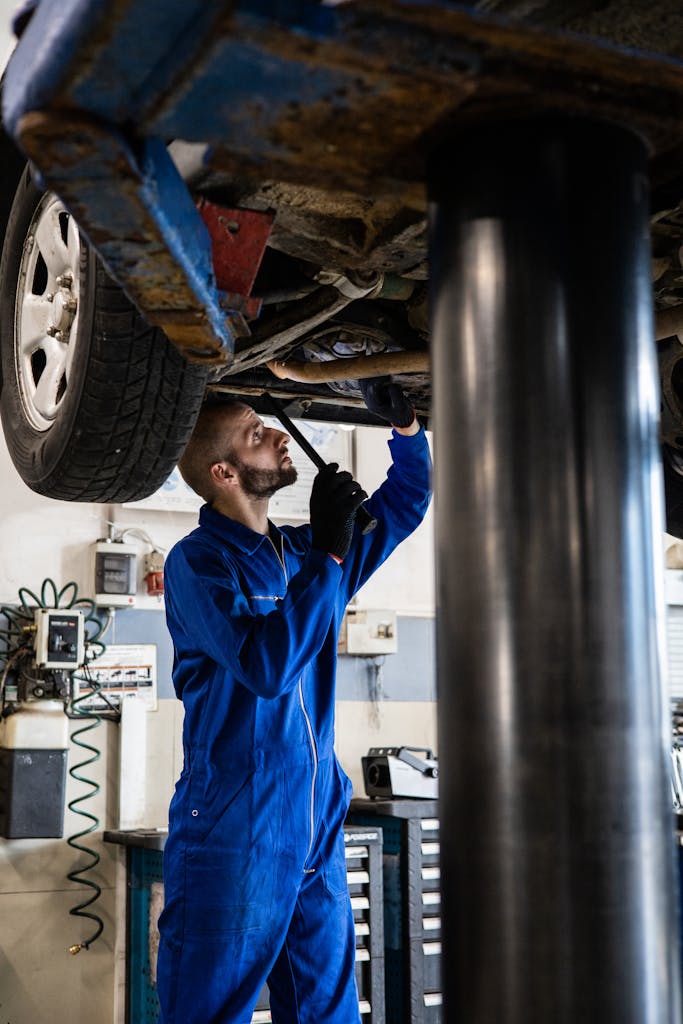 Mechanic in blue coveralls inspecting car undercarriage on hydraulic lift.