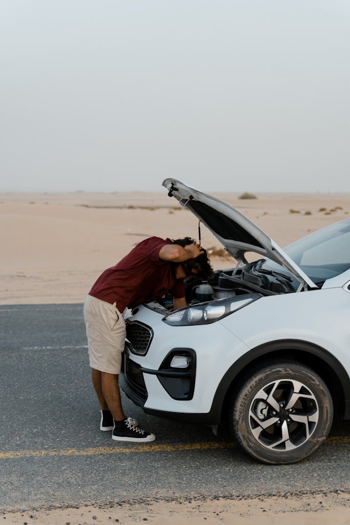 Imagen actual: A man checks under the hood of a broken car on a deserted road, surrounded by a desert landscape.