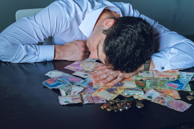 A tired businessman rests his head on a table covered with Argentine pesos and coins.