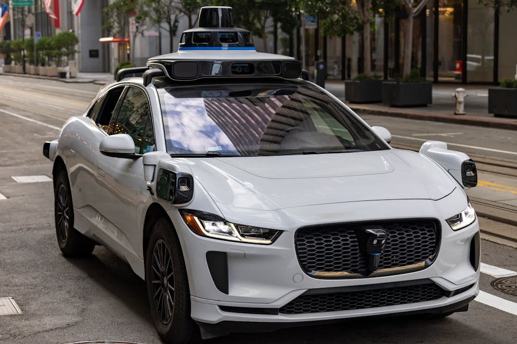 Imagen actual: A white autonomous vehicle navigating a city street, reflecting urban architecture in daylight.