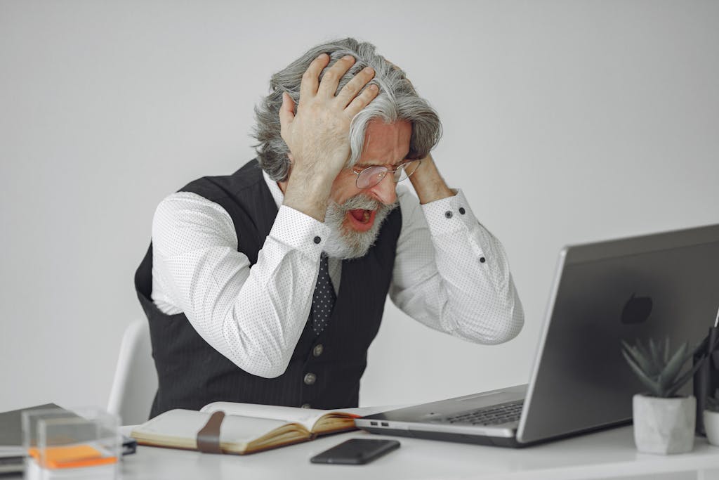 Imagen actual: Senior businessman sitting at desk, expressing frustration with his laptop in a professional office setting.