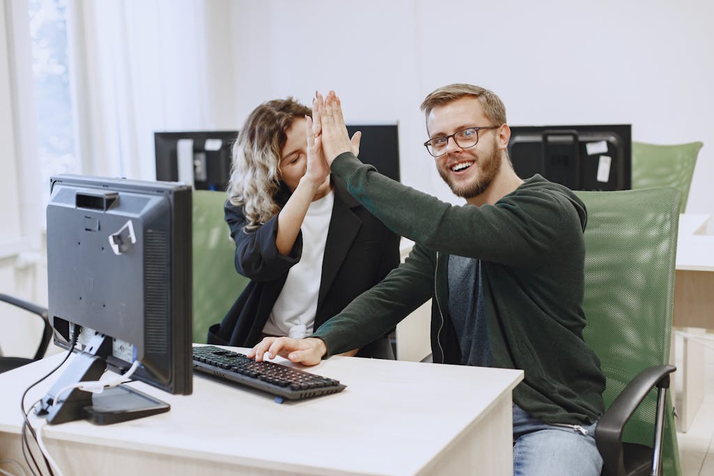 Imagen actual: Two coworkers in modern office giving a high five in front of computer screens, symbolizing teamwork.