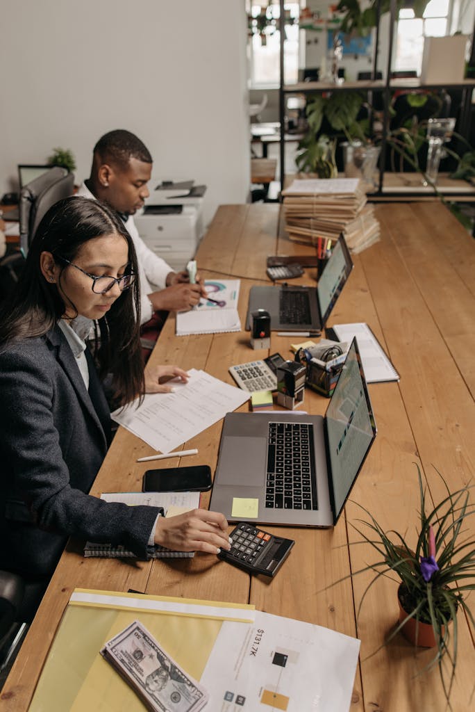 Imagen actual: Two diverse accountants working at a shared office table using laptops and calculators.