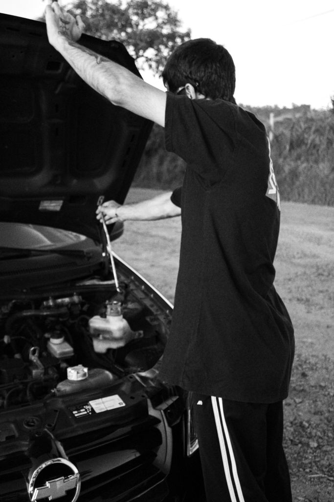 A man checks the car engine outdoors in a monochromatic setting, emphasizing mechanics and repair.