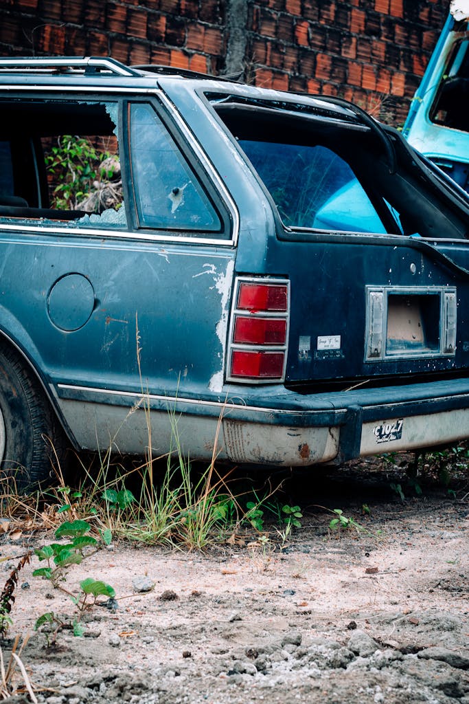 An old, abandoned station wagon in disrepair parked on rough terrain with overgrown plants.